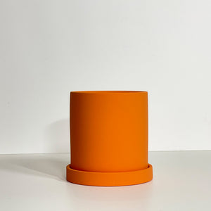 The Cyn is a bright orange cylinder-shaped planter pot. The planter pot has a drainage hole and a saucer that helps catch water.