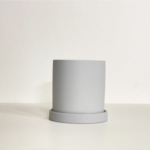 The Cyn is a grey color cylinder-shaped planter pot. The planter pot has a drainage hole and a saucer that helps catch water.