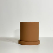 Load image into Gallery viewer, The Cyn is a brown cylinder-shaped planter pot. The planter pot has a drainage hole and a saucer that helps catch water.
