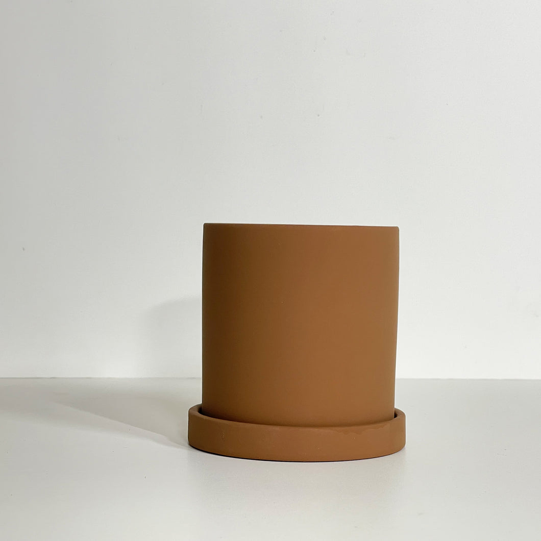 The Cyn is a brown cylinder-shaped planter pot. The planter pot has a drainage hole and a saucer that helps catch water.